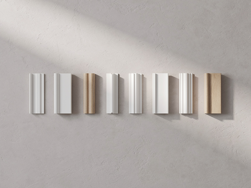 Flat lay of several molding and baseboard profiles arranged in a row on a pale plaster surface.